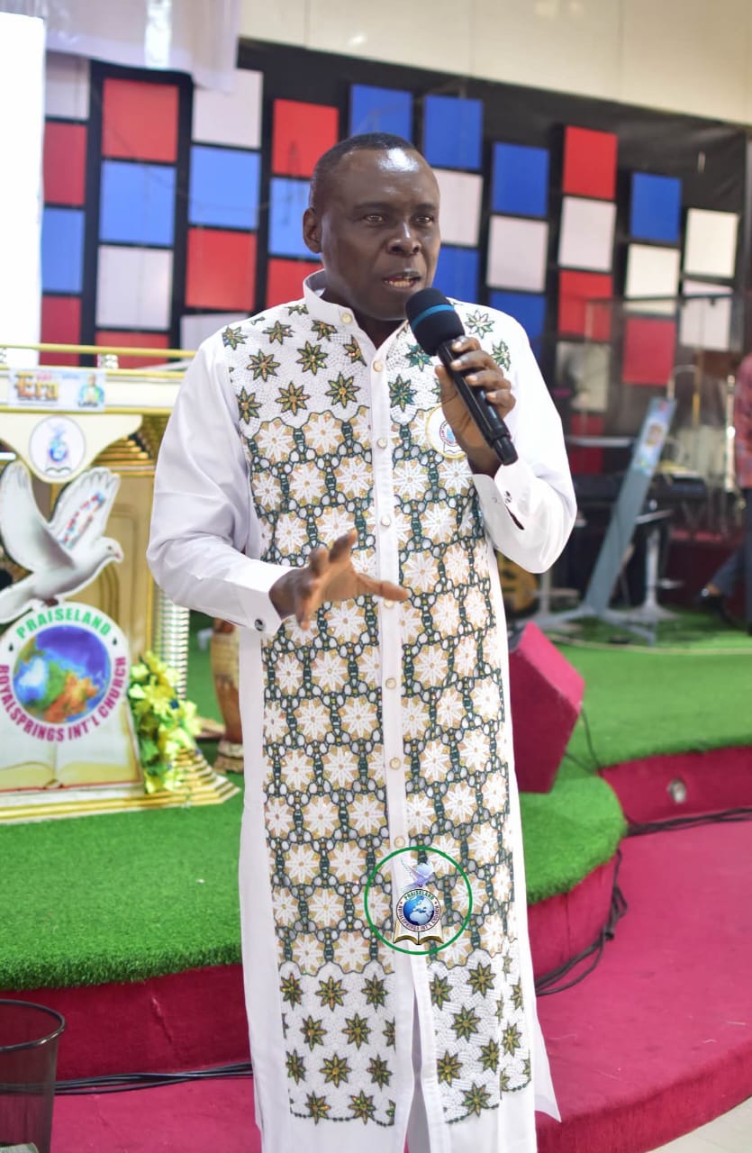Apostle Prof. E. C. Ejiofor preaching into a microphone on a green-turfed stage, wearing a white long-sleeved tunic with a green and gold floral star-patterned decorative panel, framed by a red, blue and white panelled wall