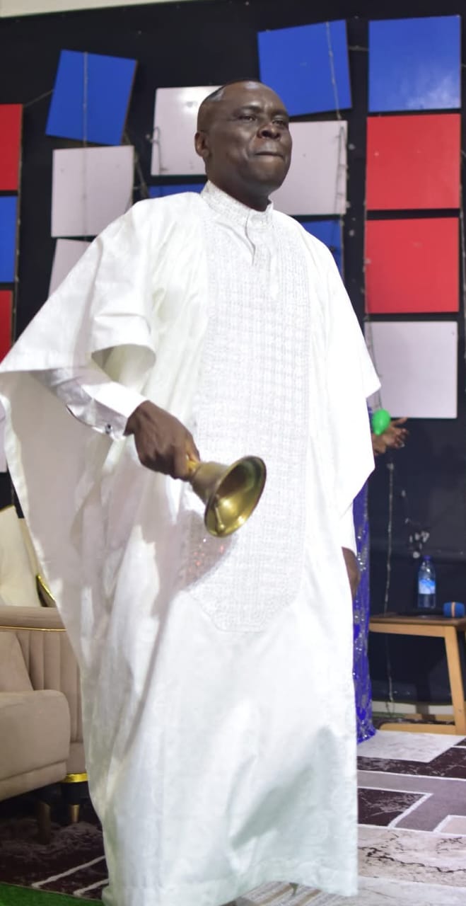 Apostle Prof. E. C. Ejiofor standing in a white Agbada garment with embroidered central panel, holding a brass hand bell in his right hand, in a studio with a dark wall of colored panels and string lights