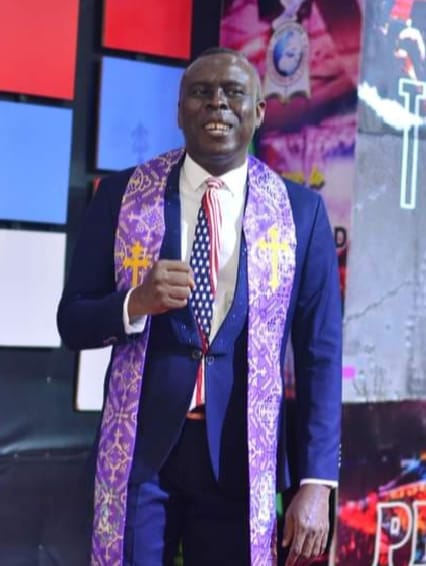 Apostle Prof. E. C. Ejiofor preaching in a navy blue suit with a purple and gold embroidered stole and an American-flag patterned tie