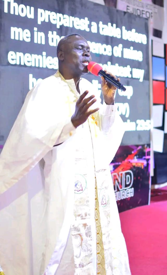 Apostle Prof. E. C. Ejiofor preaching in a white robe with ornate gold patterned lapels, holding a red microphone with an open hand extended at Praiseland Royal Springs Int'l Church