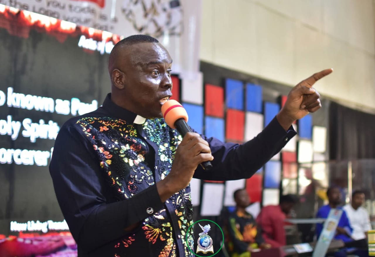 Apostle Prof. E. C. Ejiofor speaking into a microphone with his left index finger pointing, wearing a dark navy clerical shirt embellished with multi-coloured sequins and beads, with a digital screen and wall of red, blue and white panels behind him
