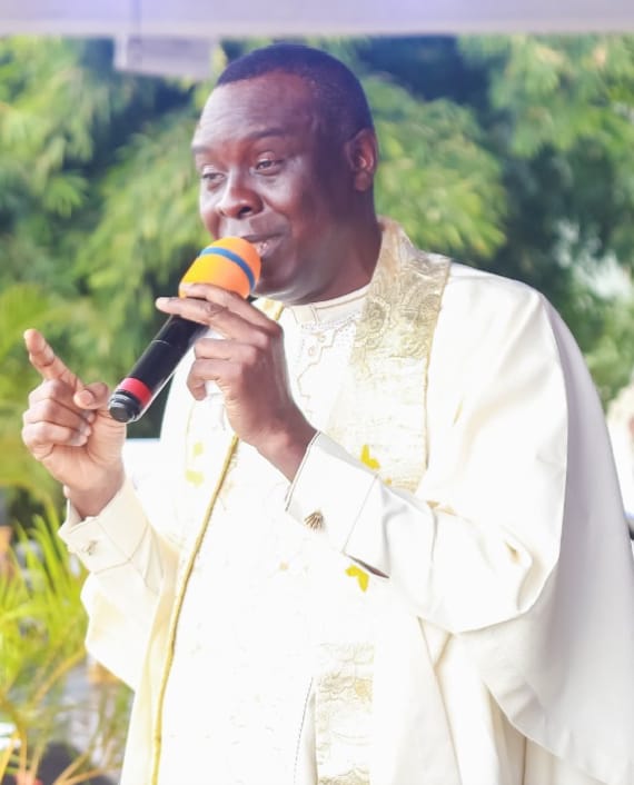 Apostle Prof. E. C. Ejiofor in a white robe with gold-embroidered stole, holding a microphone with an orange windscreen and pointing upward, outdoors with green foliage — General Overseer of Praiseland Royal Springs Int'l Church