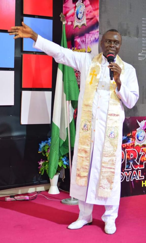 Apostle Prof. E. C. Ejiofor preaching in a white robe with ornate gold stole, holding a microphone with one arm raised at Praiseland Royal Springs Int'l Church