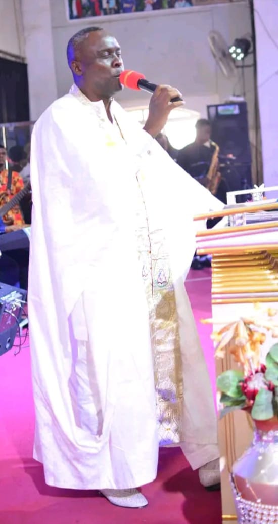 Apostle Prof. E. C. Ejiofor in a flowing white robe with a gold embroidered stole, ministering with a red microphone