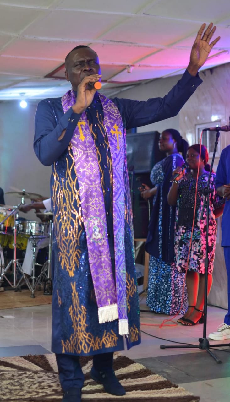 Apostle Prof. E. C. Ejiofor ministering in worship with a raised hand, wearing a navy robe and purple stole