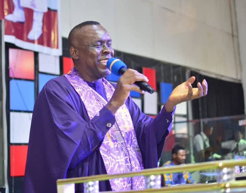 Apostle Prof. E. C. Ejiofor preaching in a purple robe with an embroidered stole, holding a microphone at Praiseland Royal Springs Int'l Church