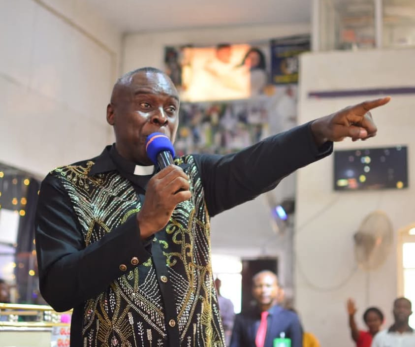 Apostle Prof. E. C. Ejiofor in a black clerical shirt with gold embroidery, preaching and pointing