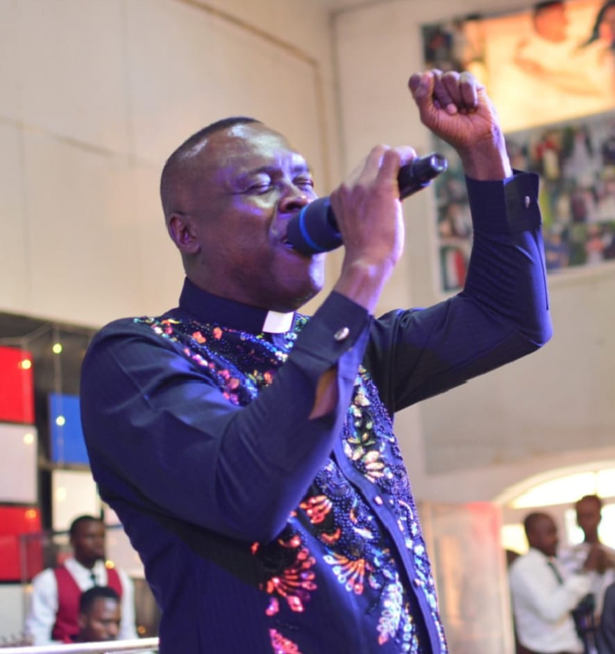 Apostle Prof. E. C. Ejiofor singing into a microphone with his left fist raised, wearing a navy blue clerical shirt with a white collar tab and colourful sequined floral patterns, in a church setting with a red, white and blue panelled wall behind him