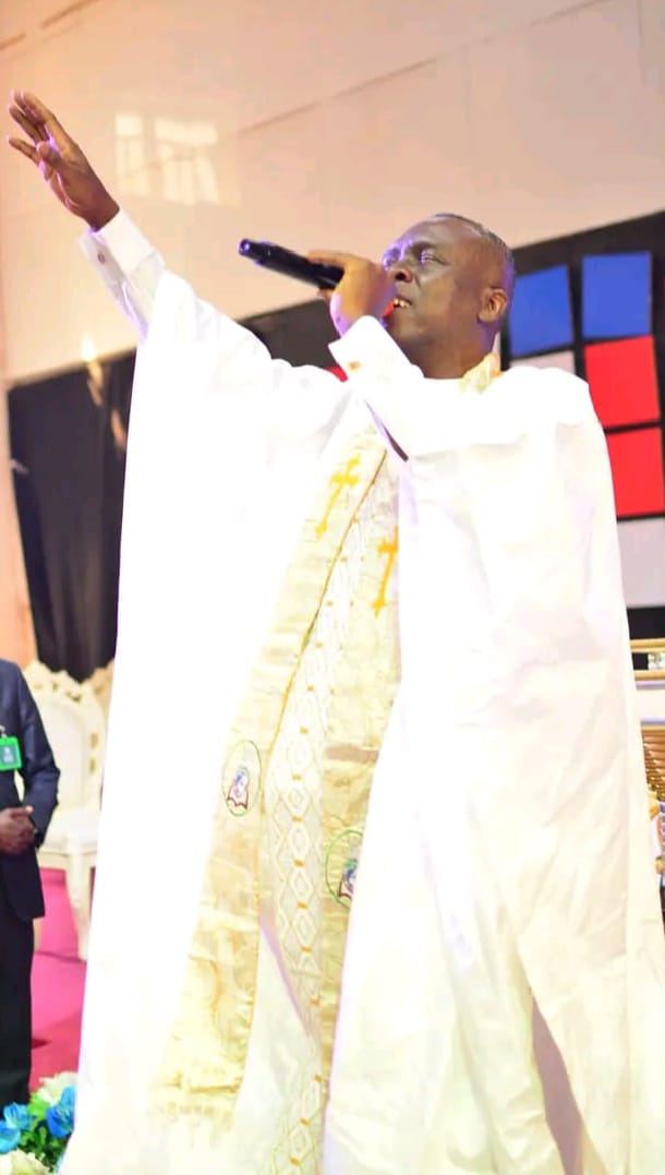 Apostle Prof. E. C. Ejiofor wearing a bright white robe with a gold-patterned stole, holding a microphone to his mouth with his left hand and raising his right hand in worship, standing on a church stage with red and blue wall panels behind him