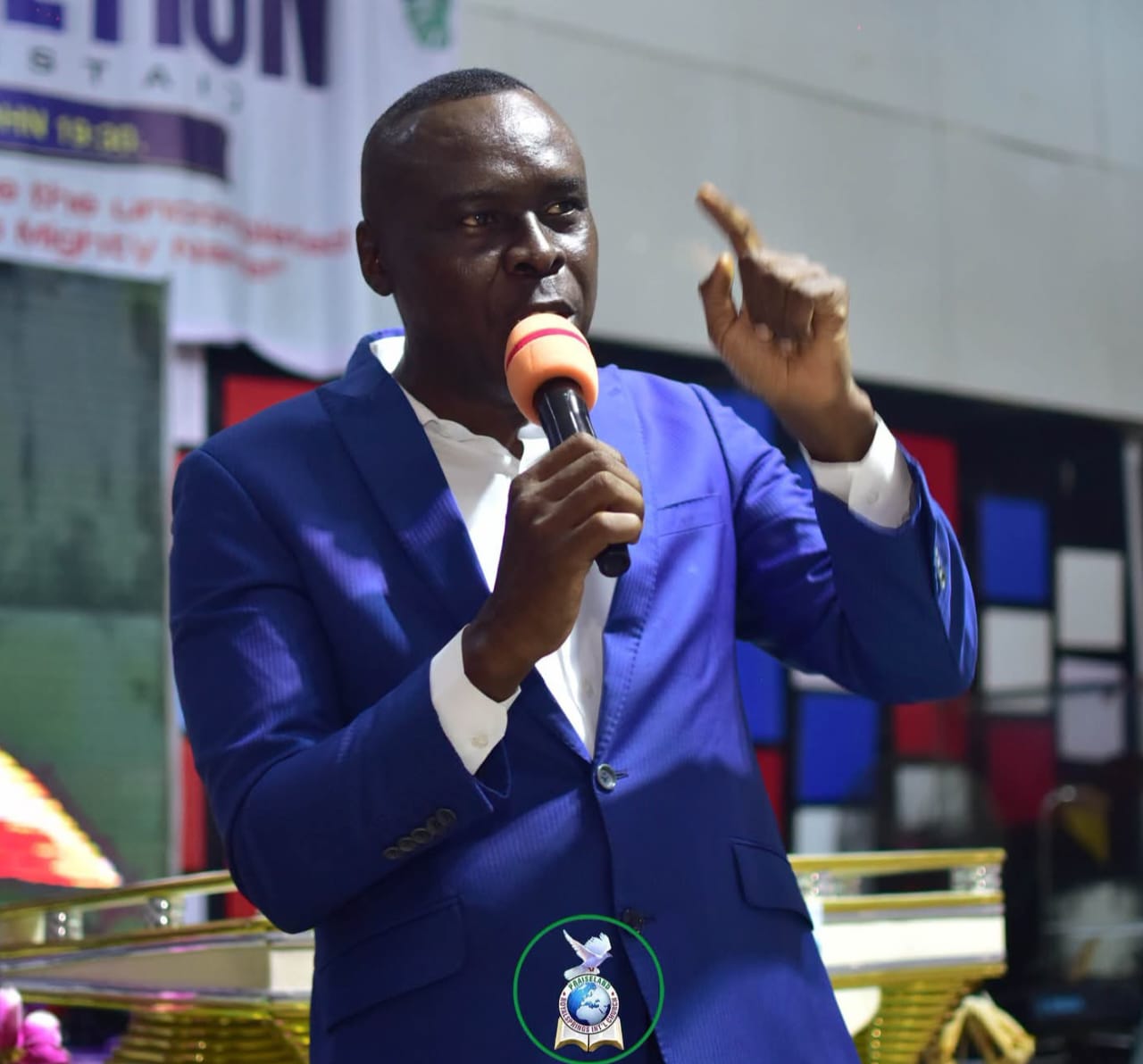Apostle Prof. E. C. Ejiofor speaking into a microphone with his left index finger pointing upward, wearing a royal blue blazer over a white dress shirt, standing behind a gold podium on a church stage with a wall of red, blue and white panels behind him
