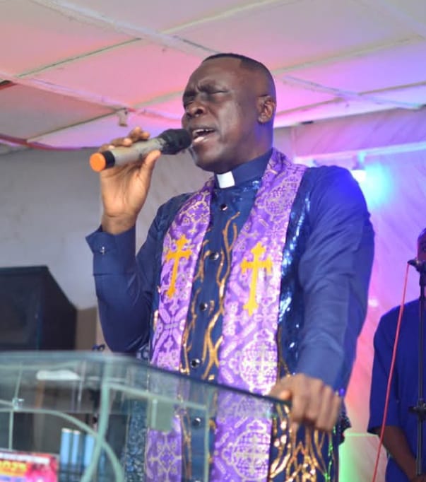 Apostle Prof. E. C. Ejiofor in a blue clerical shirt and purple stole with gold crosses, praying into a microphone at the pulpit