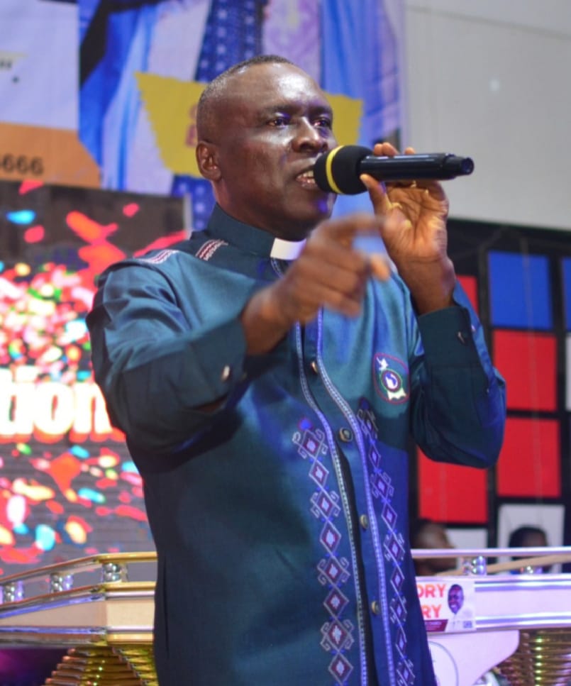Apostle Prof. E. C. Ejiofor in a teal clerical robe, microphone in hand, pointing toward the congregation