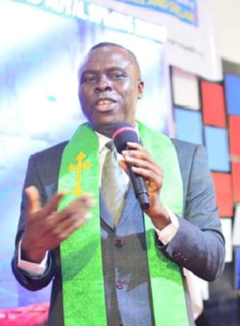 Apostle Prof. E. C. Ejiofor preaching with a microphone, wearing a grey suit and a green stole with a gold cross
