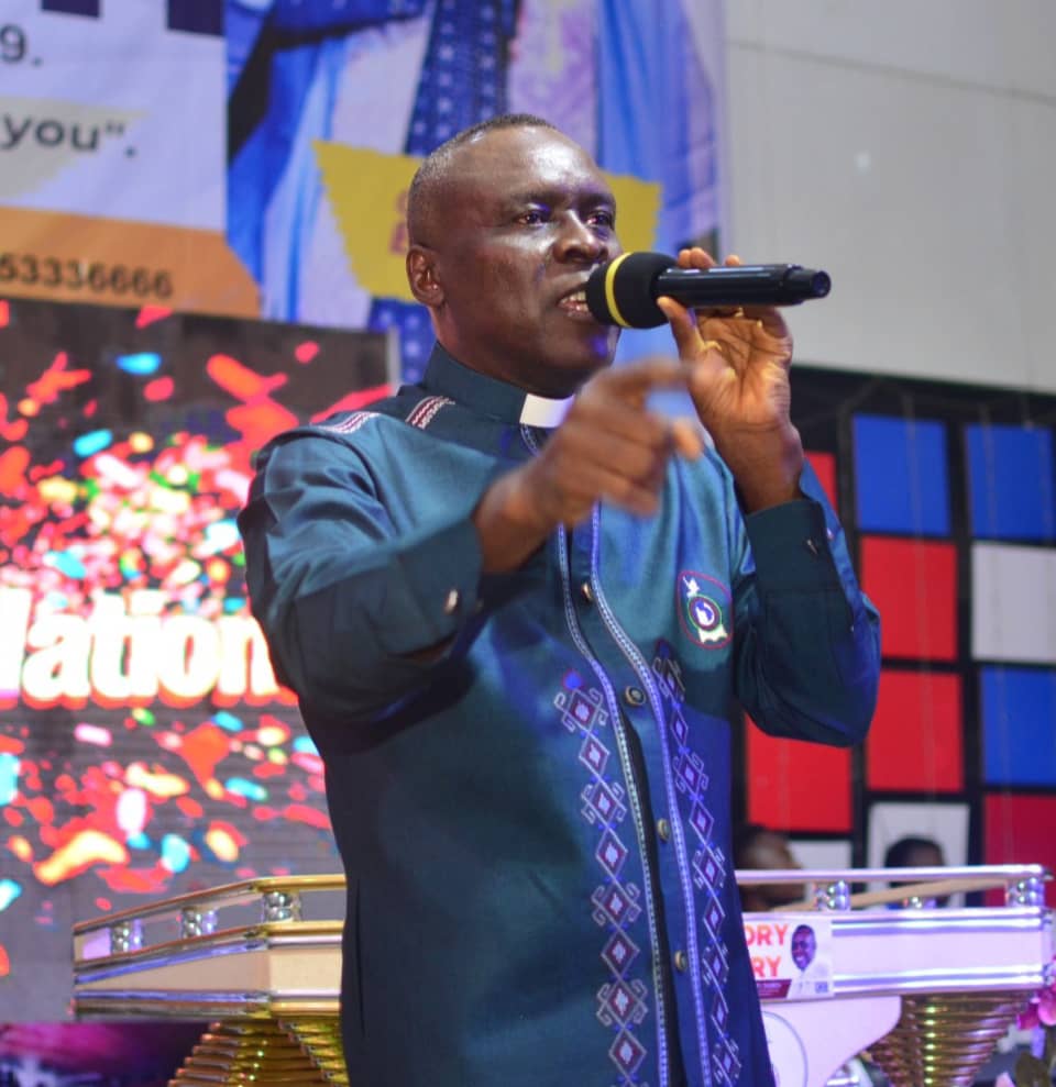 Apostle Prof. E. C. Ejiofor preaching in a teal embroidered robe, holding a microphone at Praiseland Royal Springs Int'l Church