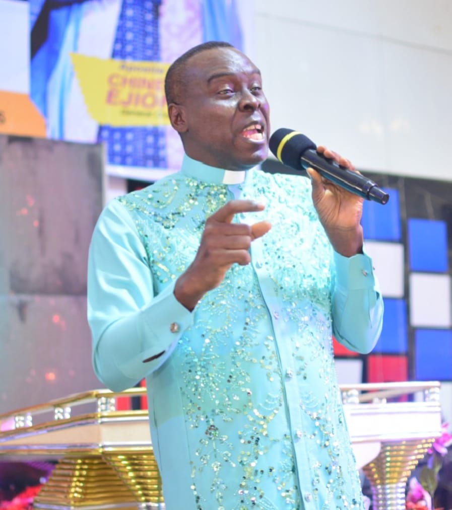 Apostle Prof. E. C. Ejiofor wearing a turquoise sequin-embellished clerical shirt with a white collar, speaking into a microphone with a podium with gold supports before him, on a church stage with blue, white and red background panels