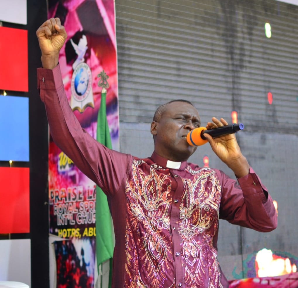 Apostle Prof. E. C. Ejiofor ministering with a raised fist and microphone in a maroon embroidered outfit at an open-air service