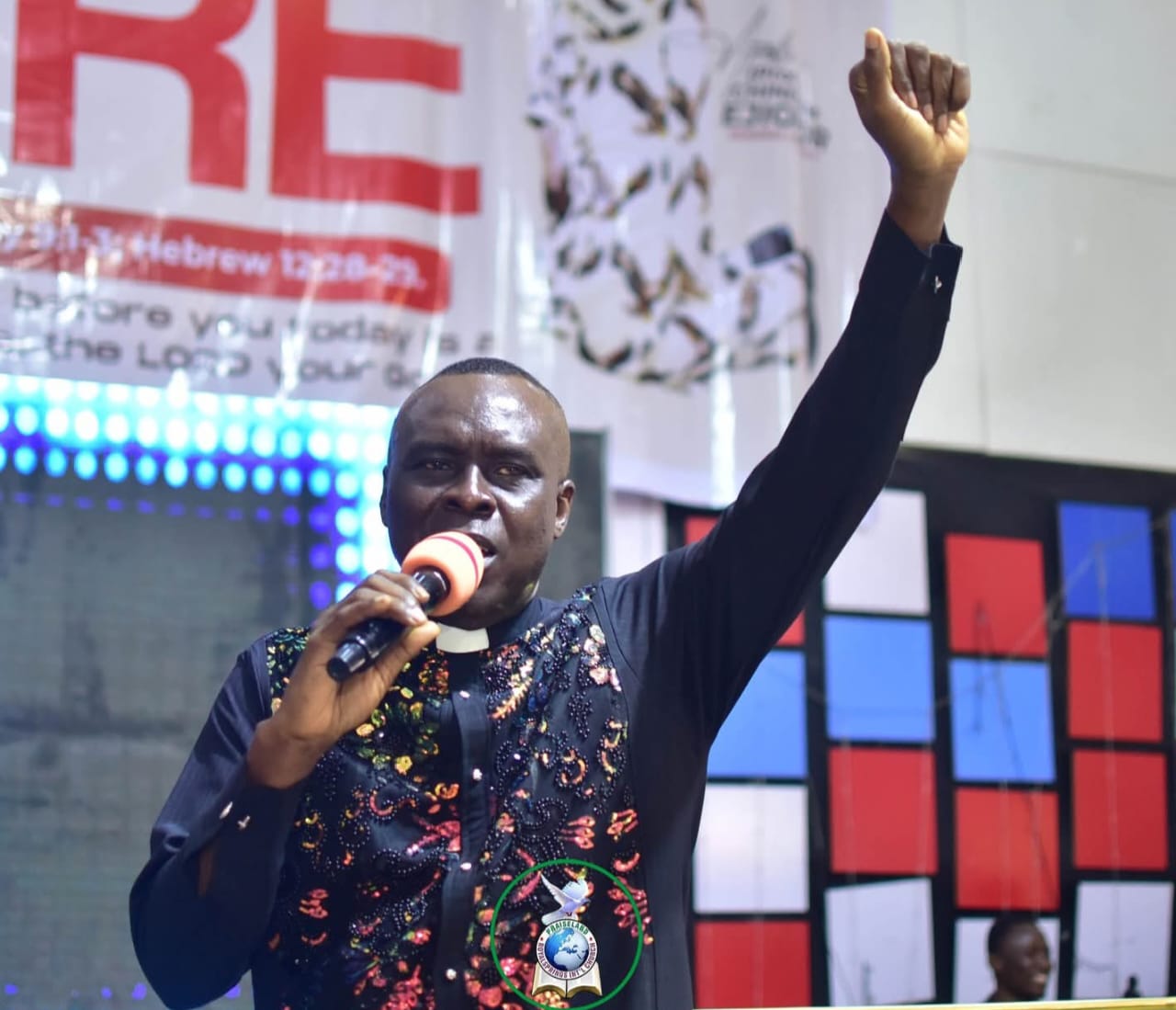 Apostle Prof. E. C. Ejiofor in a sequined black clerical shirt with raised fist, ministering with a microphone at Praiseland Royal Springs Int'l Church