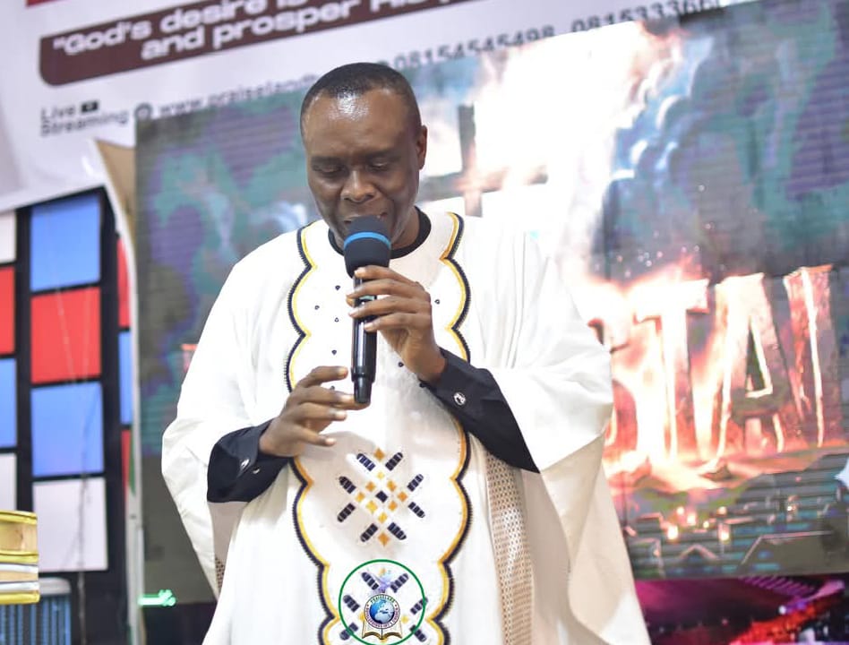 Apostle Prof. E. C. Ejiofor in a white embroidered ceremonial robe, speaking into a microphone on the church stage with a colourful backdrop
