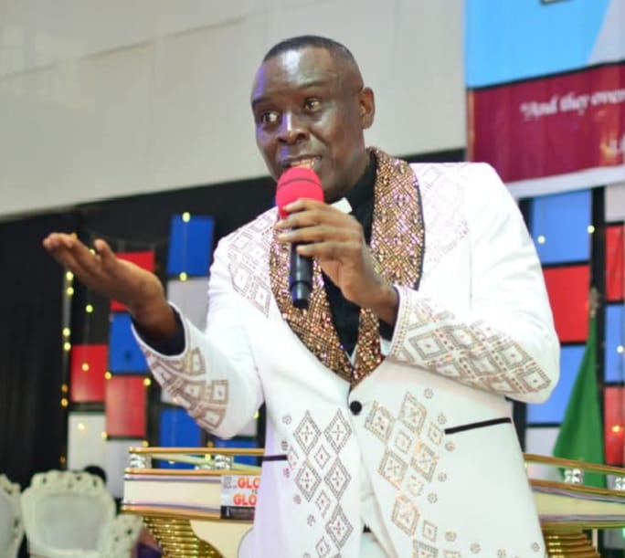 Apostle Prof. E. C. Ejiofor preaching in a white suit with ornate gold patterned lapels, holding a red microphone with an open hand extended at Praiseland Royal Springs Int'l Church