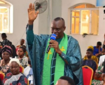 Apostle Prof. E. C. Ejiofor in a green robe with a cross-embroidered stole, hand raised, speaking into a blue microphone among the congregation