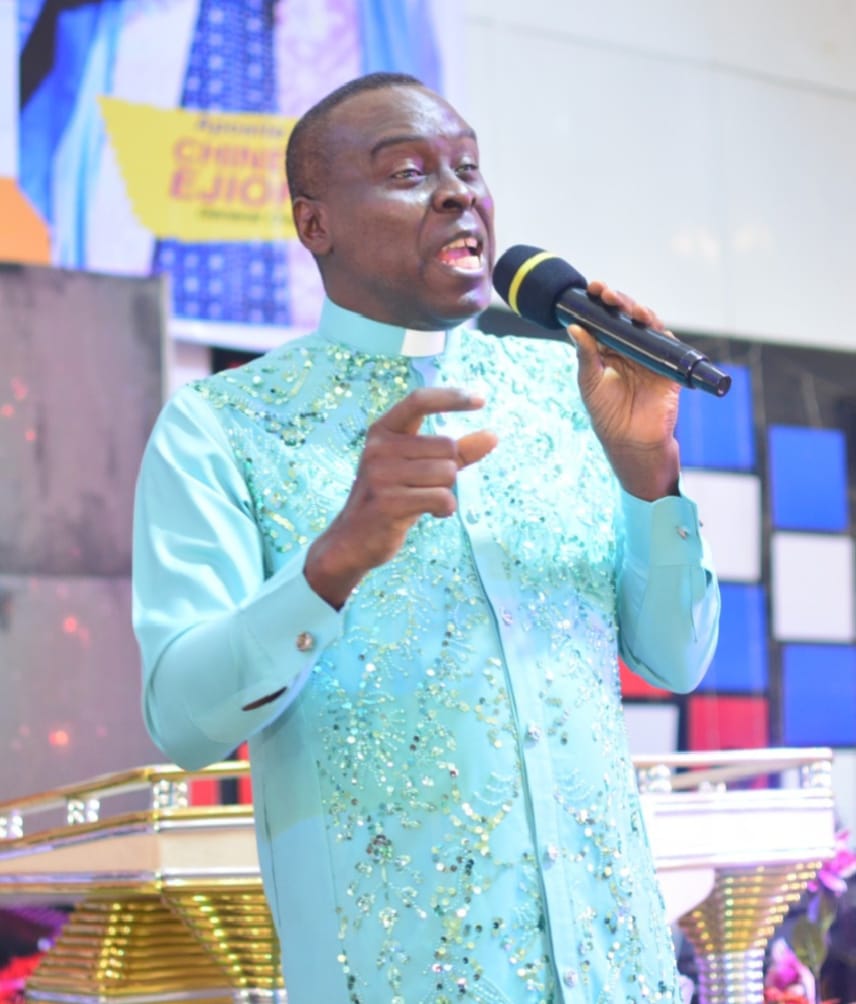 Apostle Prof. E. C. Ejiofor preaching in a light blue sequined robe, holding a microphone and pointing, at Praiseland Royal Springs Int'l Church