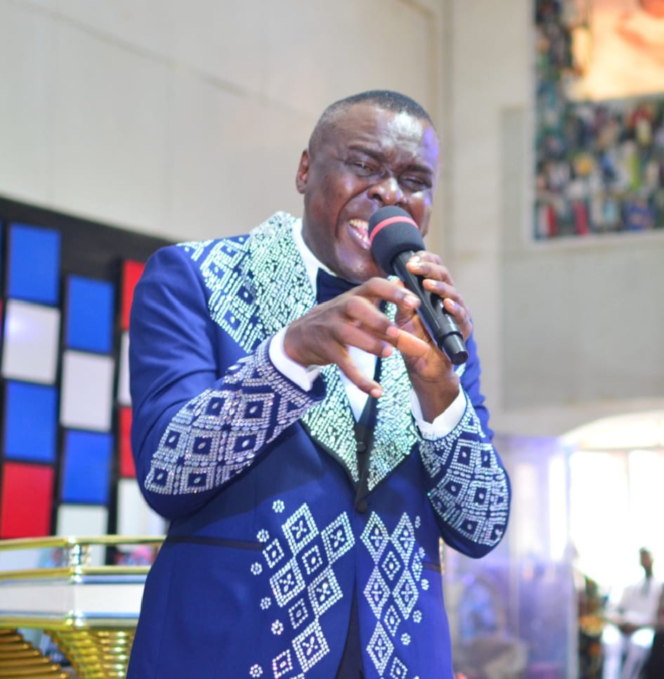 Apostle Prof. E. C. Ejiofor preaching with joyful fervour on a church stage, wearing a royal blue blazer with silver rhinestone embellishment and holding a microphone — General Overseer of Praiseland Royal Springs Int'l Church