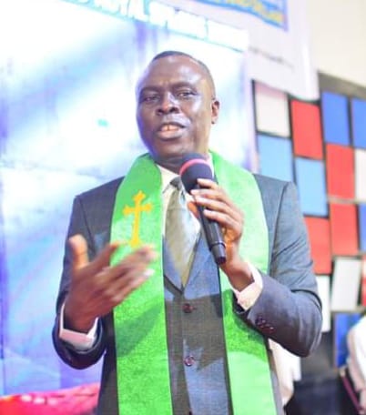 Apostle Prof. E. C. Ejiofor in a grey suit and green stole with a gold cross, preaching with a microphone