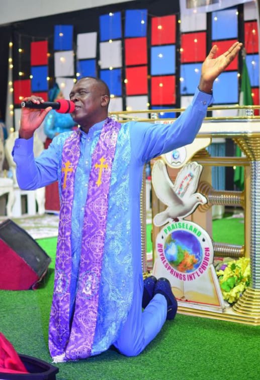 Apostle Prof. E. C. Ejiofor kneeling in prayer in a blue robe with purple stole at Praiseland Royal Springs Int'l Church