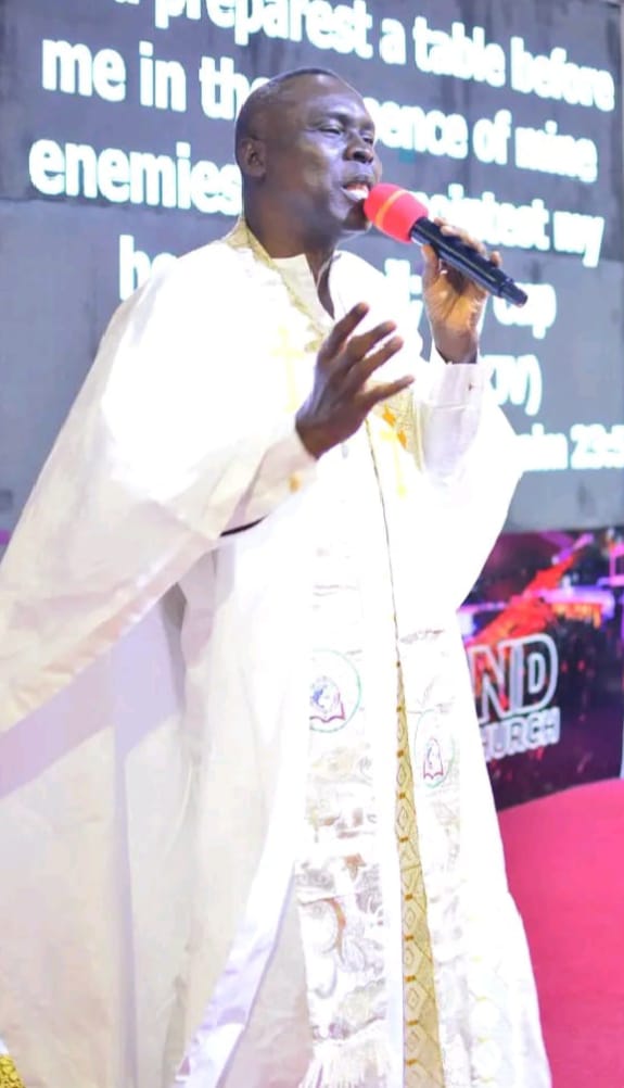 Apostle Prof. E. C. Ejiofor in white liturgical vestments with a gold and silver embroidered stole, speaking into a microphone on a church stage with a Bible verse from Psalm 23:5 displayed on the background screen