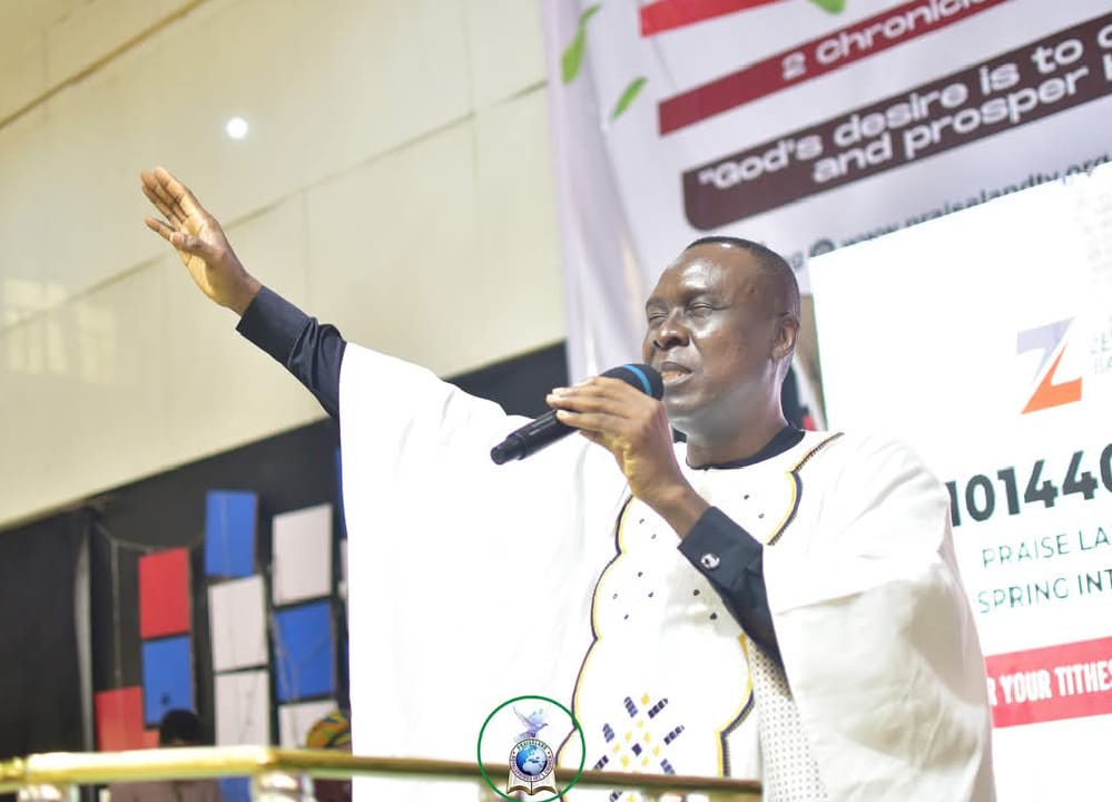Apostle Prof. E. C. Ejiofor preaching into a microphone with his right arm raised and palm open, wearing a flowing white Agbada garment with a central embroidered panel in gold, black and blue, on a church stage with colored panel backdrop