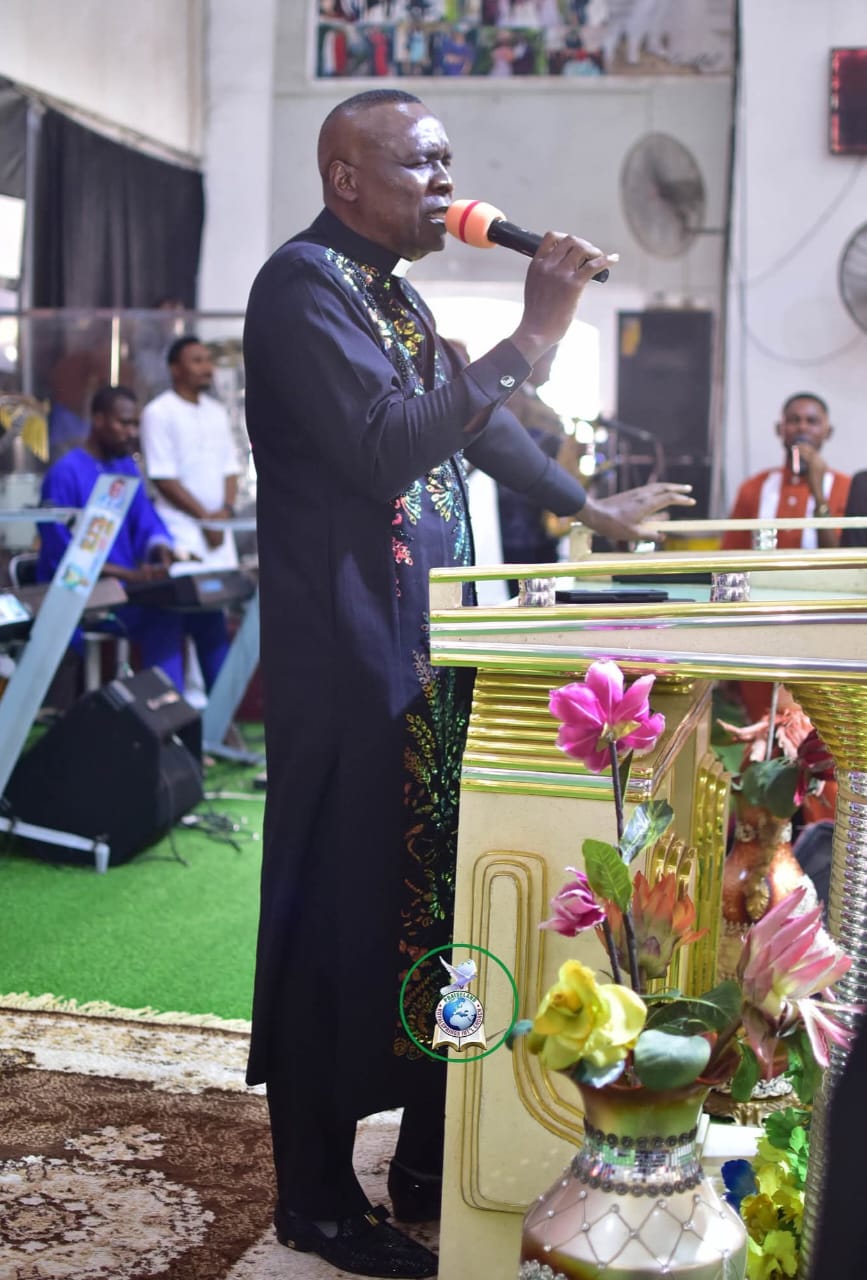Apostle Prof. E. C. Ejiofor wearing a black clerical robe with a white collar and shimmering multi-colored sequin embroidery, holding a microphone to his mouth behind an ornate gold pulpit on a church stage