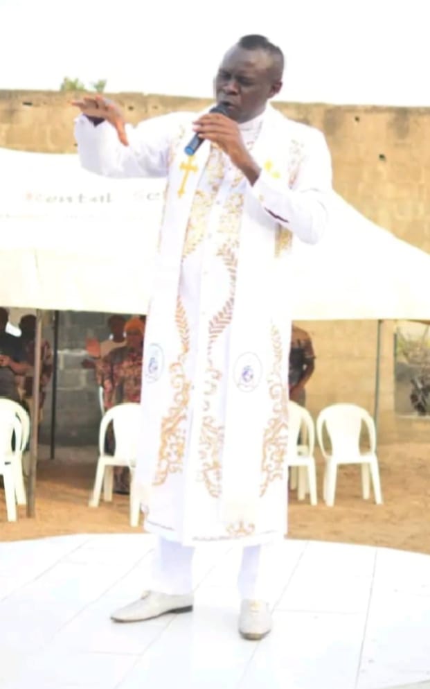 Apostle Prof. E. C. Ejiofor in a white and gold clerical robe, ministering with a microphone at an outdoor service