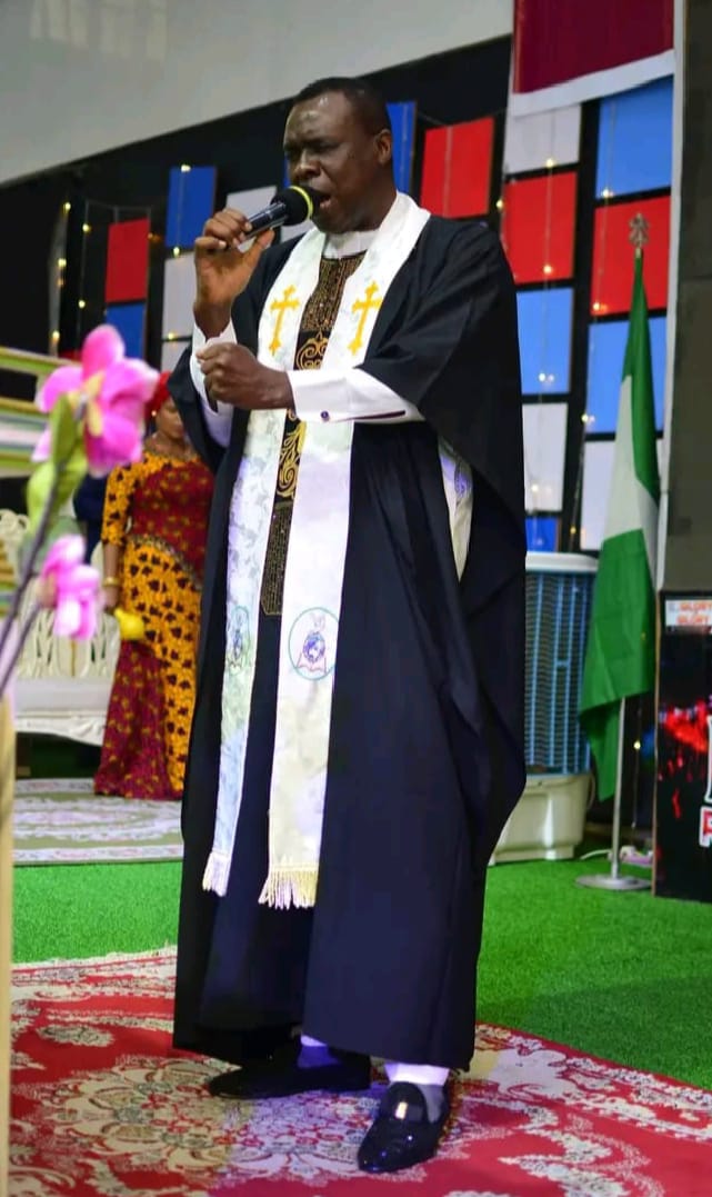 Apostle Prof. E. C. Ejiofor preaching in a black robe with white and gold embroidered stole, holding a microphone at Praiseland Royal Springs Int'l Church