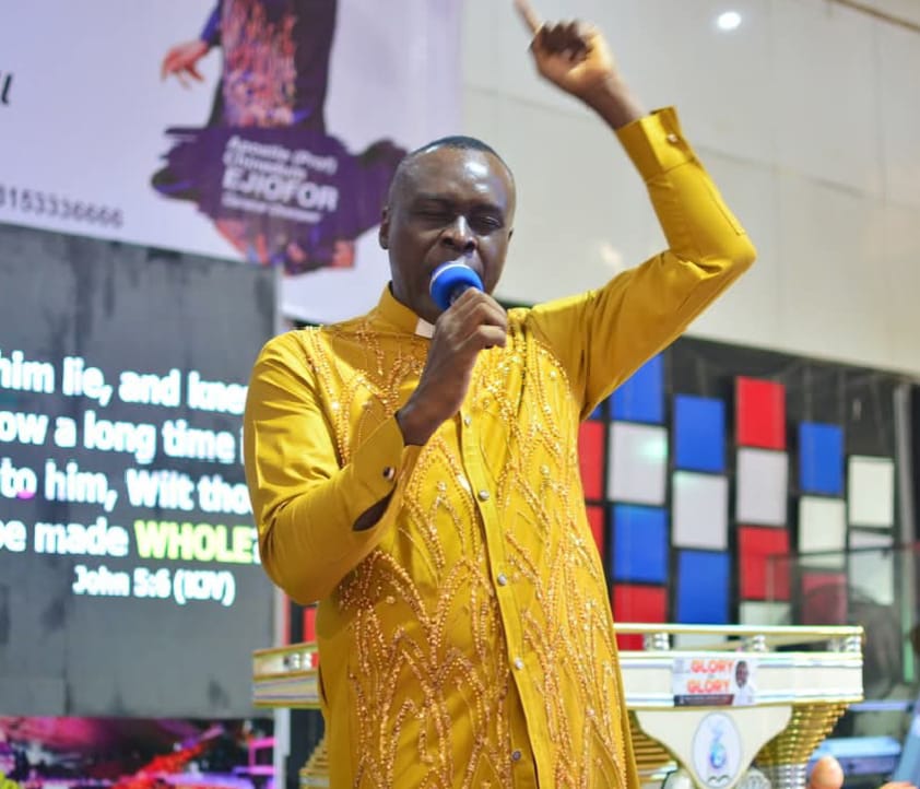 Apostle Prof. E. C. Ejiofor in a gold embroidered clerical outfit, preaching into a blue microphone with his hand raised