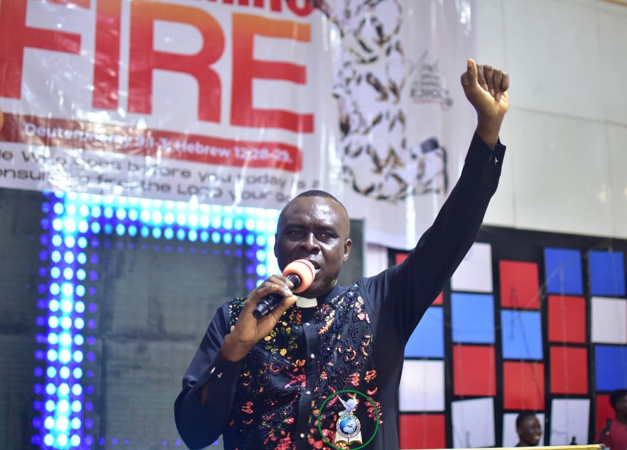 Apostle Prof. E. C. Ejiofor preaching with a microphone, one arm raised in a fist, wearing a black clerical shirt with colourful embroidered patterns