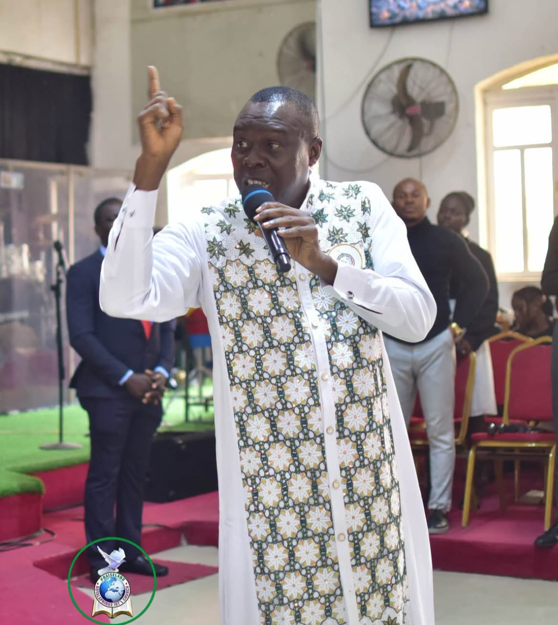 Apostle Prof. E. C. Ejiofor preaching into a microphone with his index finger raised, wearing a white tunic with a green and gold floral star-patterned decorative panel, on a green-turfed stage with red-carpeted steps