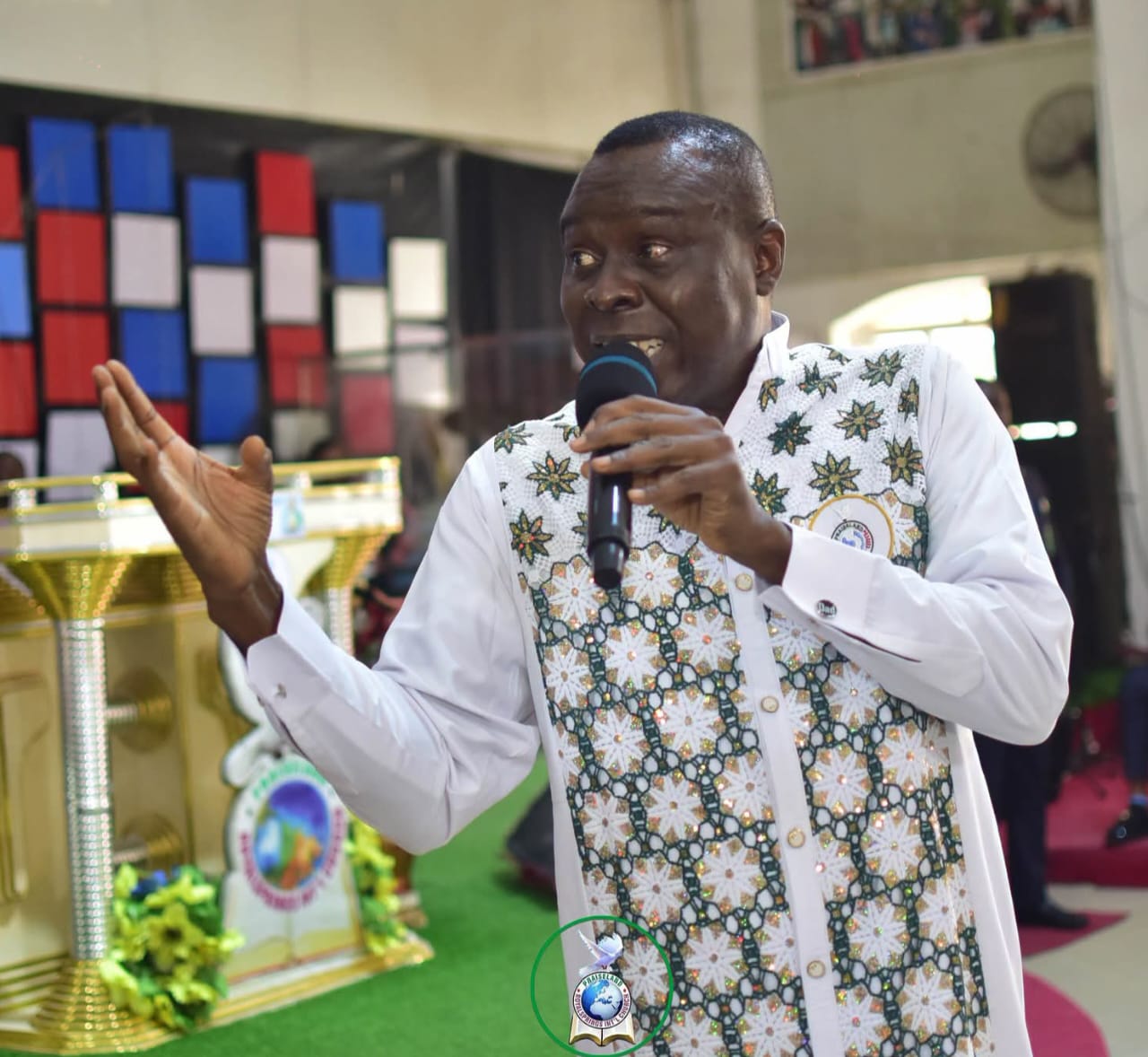 Apostle Prof. E. C. Ejiofor speaking into a microphone with his left hand raised in an open gesture, wearing an elaborate white tunic decorated with gold-centred star motifs and green accents, standing on a green turf floor beside an ornate gold and silver podium in a church setting